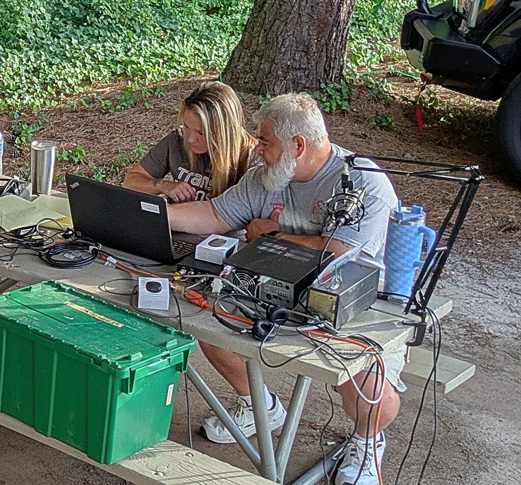 Two DeKalb ARES operators at a picnic table under tree cover, working an HF station with a laptop logging program, a transceiver and power supply, and a boom microphone, with a green equipment tote on the bench.