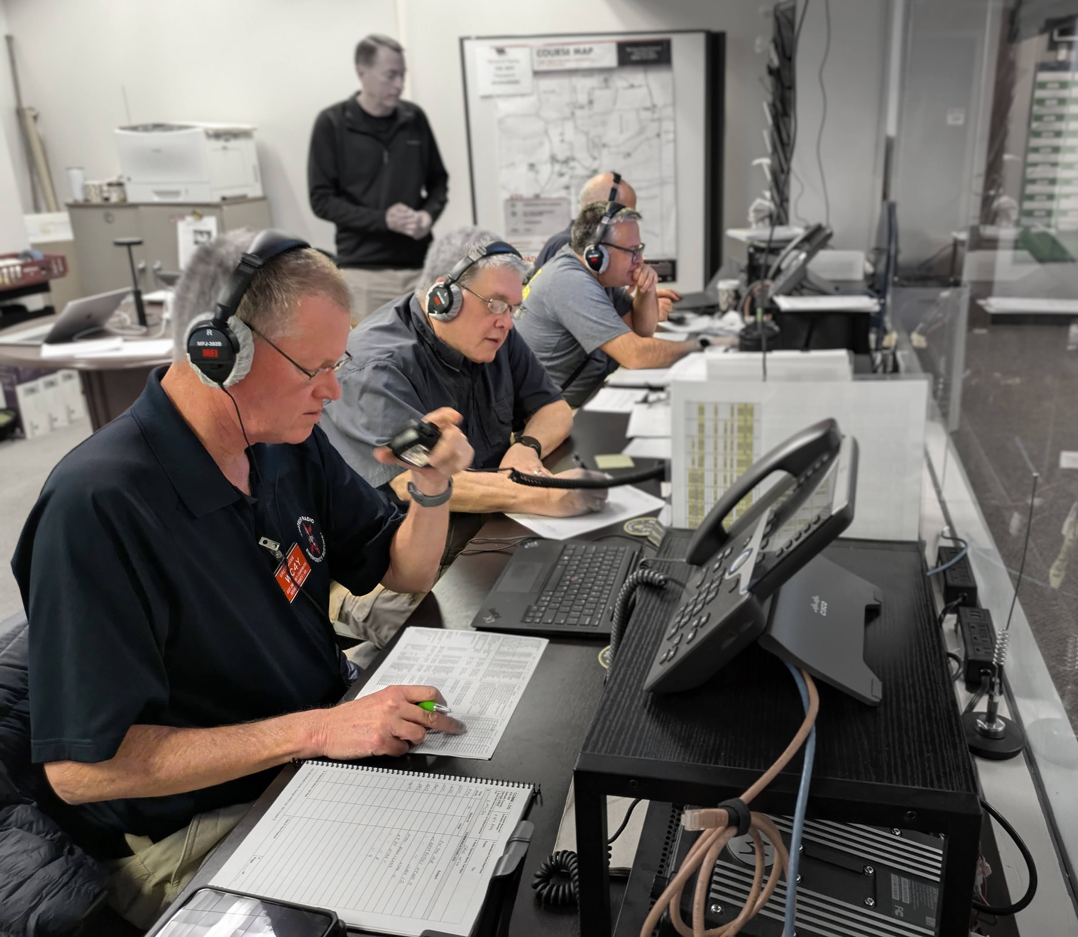 DKARES operators at the Atlanta Marathon Net Control desk — three operators in headphones, one keying a microphone, with the marathon course map on the wall behind them and paper comm logs on the desk.