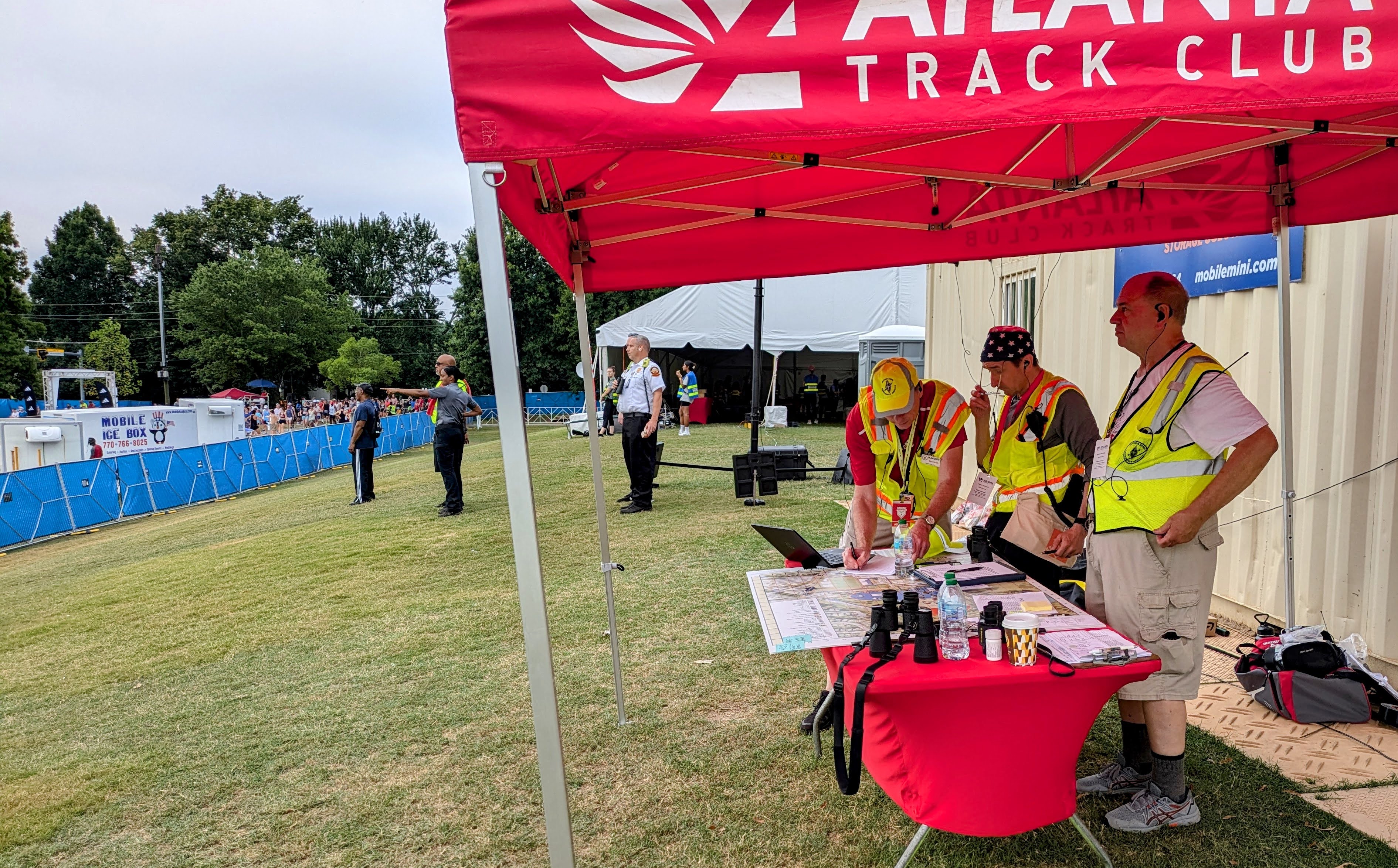 DKARES operators in safety vests at an Atlanta Track Club tent near the Peachtree Road Race finish line, working with maps, binoculars, and HT radios while runners and finish-line staff are visible behind the safety barrier.