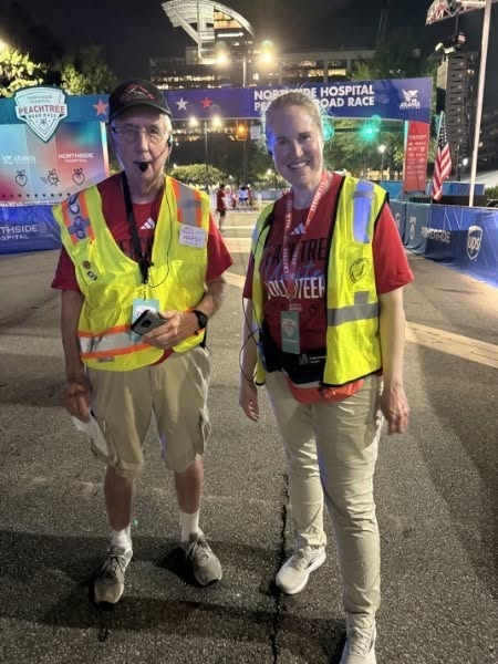 Two DeKalb ARES volunteers in yellow high-visibility safety vests stand on the course before dawn at the Peachtree Road Race finish area, in front of the illuminated Northside Hospital Peachtree Road Race finish gantry with the stadium and American flags behind them. One holds a handheld radio.
