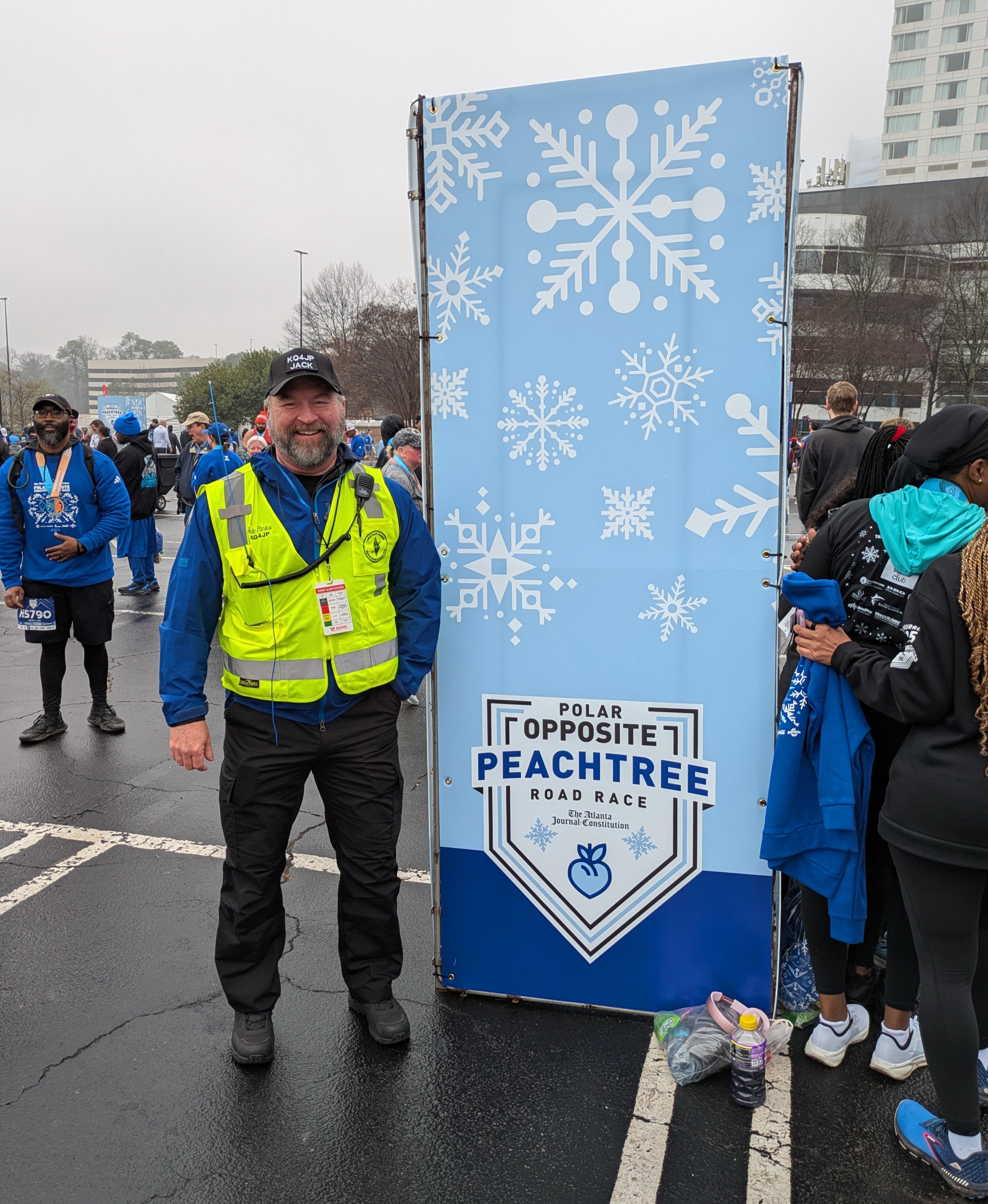 DKARES operator Jack Parks (KQ4JP) in a yellow safety vest with an HT mic on his shoulder, standing next to a tall Polar Opposite Peachtree Road Race banner at the Lenox Square finish area on a cold, foggy morning.