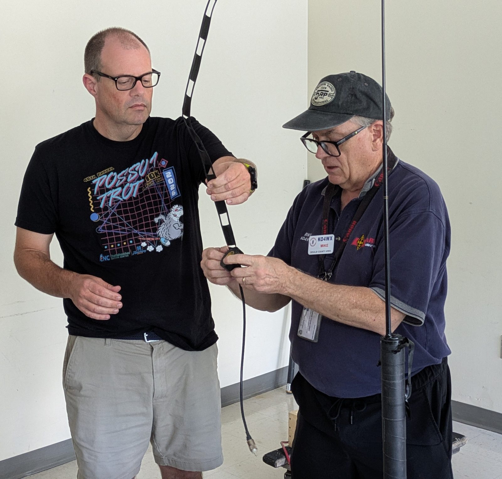 Michael Boatright (KO4WX) at the July DeKalb ARES meeting, holding the feedpoint and coax of a roll-up slim jim antenna while another member holds the black window-line element upright. A telescoping mast stands beside them.