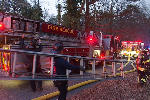 DeKalb Fire & Rescue Engine 18 ladder truck on scene at dusk, firefighters on the side rails and hose lines deployed across the foreground.