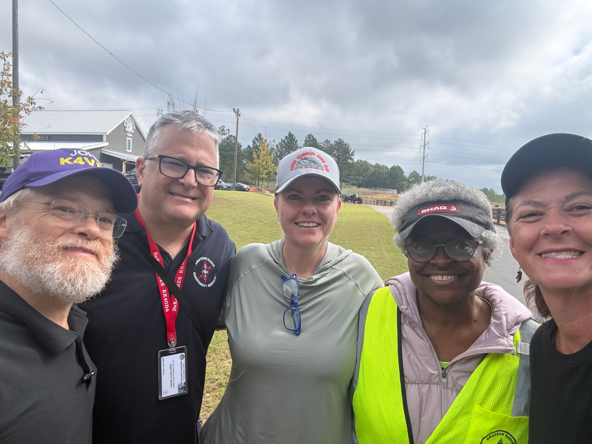 Five DeKalb ARES members standing together outdoors on an overcast day, smiling at the camera, several wearing high-visibility safety vests and radio club caps.