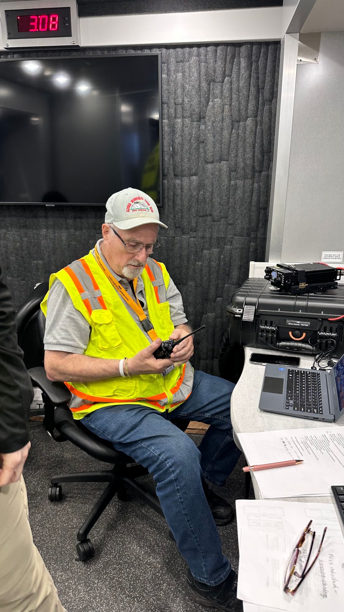 An operator in a high-visibility vest working a handheld radio at a desk inside the mobile command vehicle, with an HF radio case and laptop beside him