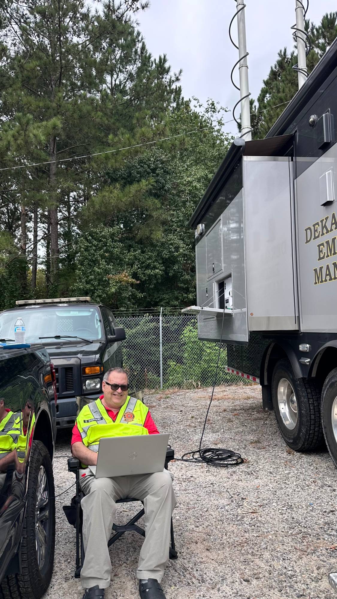 A DeKalb ARES member in a high-visibility vest sitting outdoors with a laptop beside the mobile command vehicle, cable running to the vehicle