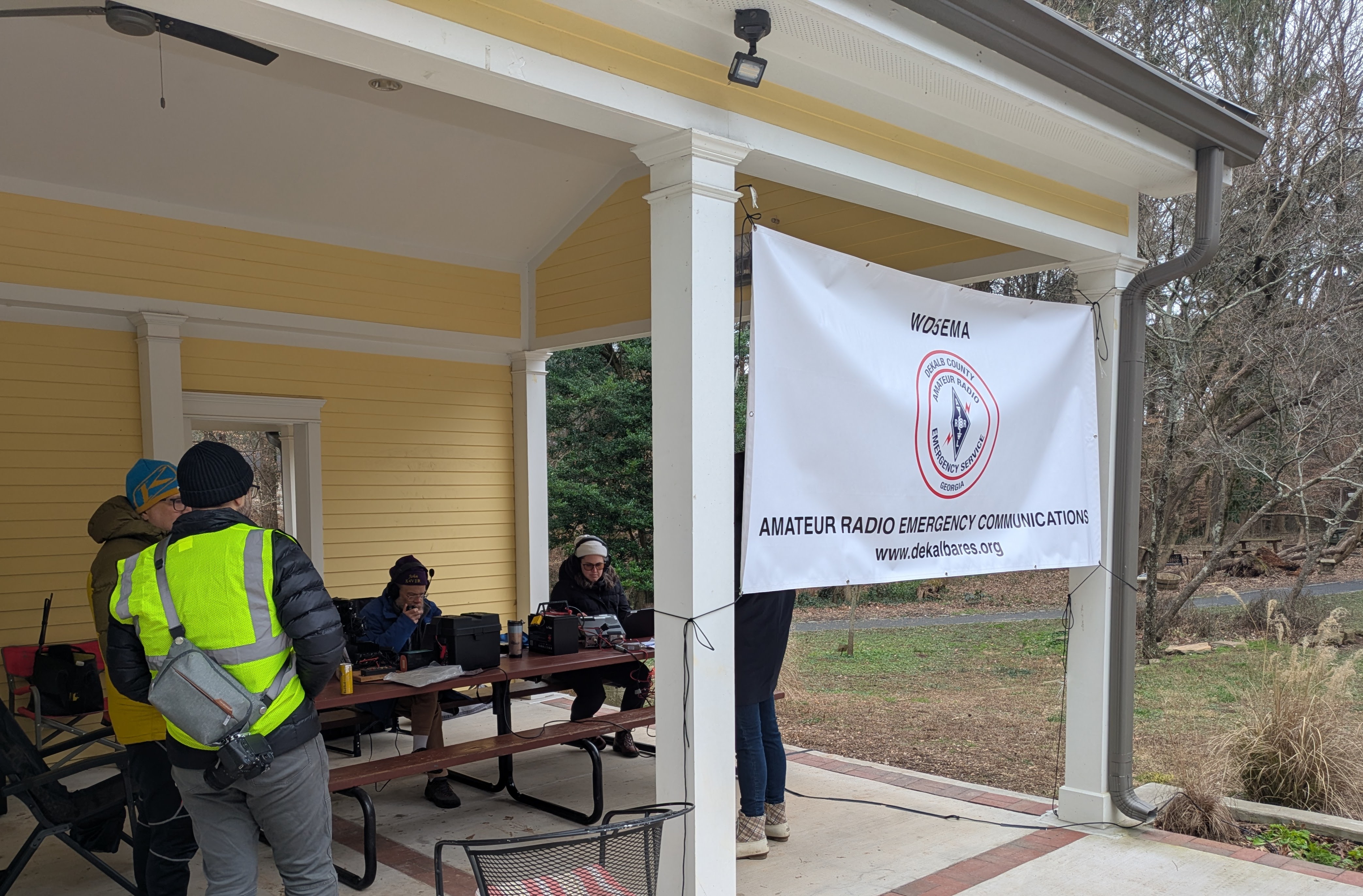 DKARES members operating Winter Field Day under a covered pavilion at Briarlake Forest Park, with the WD5EMA Amateur Radio Emergency Service banner hanging from the eaves and operators at picnic tables with HF and VHF gear.